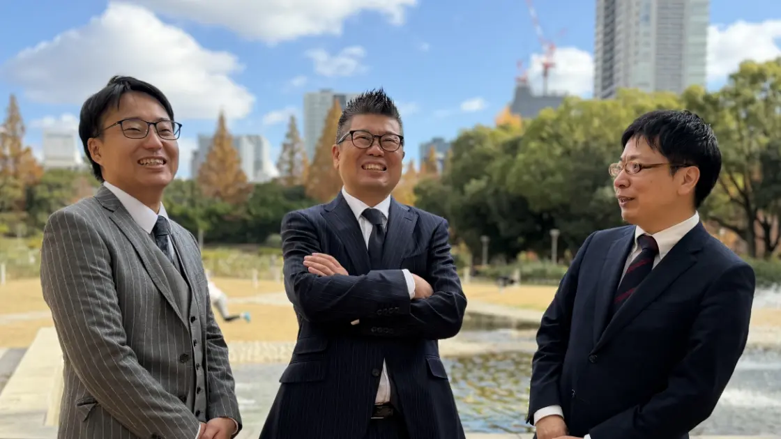 青空の下、公園の噴水を背景に笑顔で並ぶスーツ姿の男性3人。弁護士事務所のスタッフの集合写真