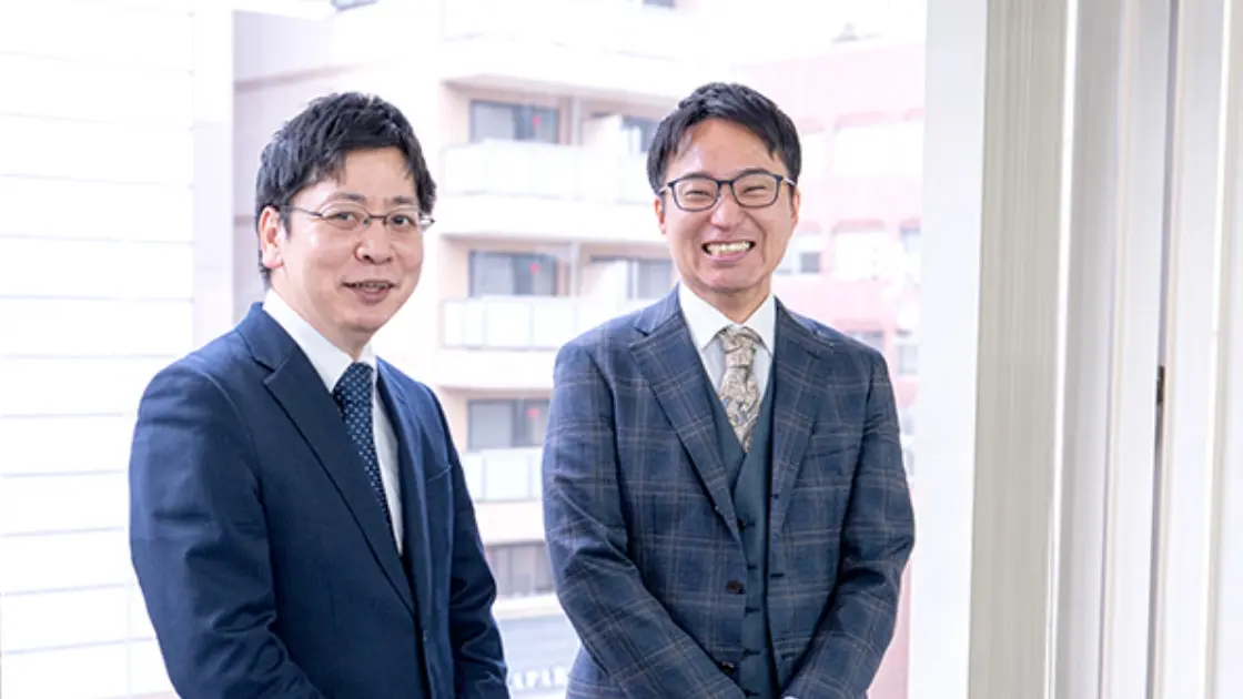 窓際で並んで笑顔を見せる、スーツ姿の二人の男性ビジネスマン