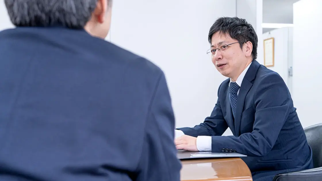 オフィス内での打ち合わせ風景。デスク越しに向かい合い、スーツ姿で話をしている様子