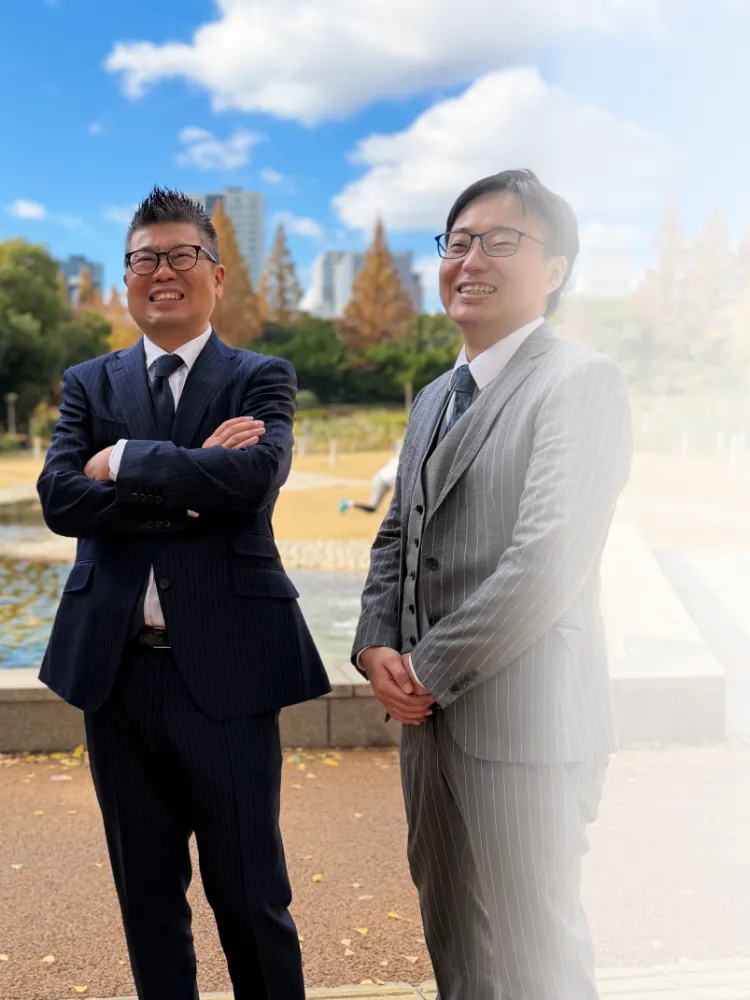 青空の下、公園の噴水を背景に笑顔で並ぶスーツ姿の男性3人。弁護士事務所のスタッフの集合写真