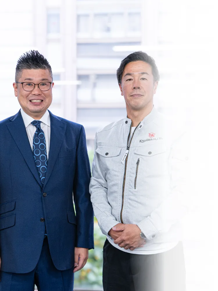 室内で並んで立つ二人の男性。左側はスーツ姿の男性、右側はロゴ入りの作業着を着用した男性