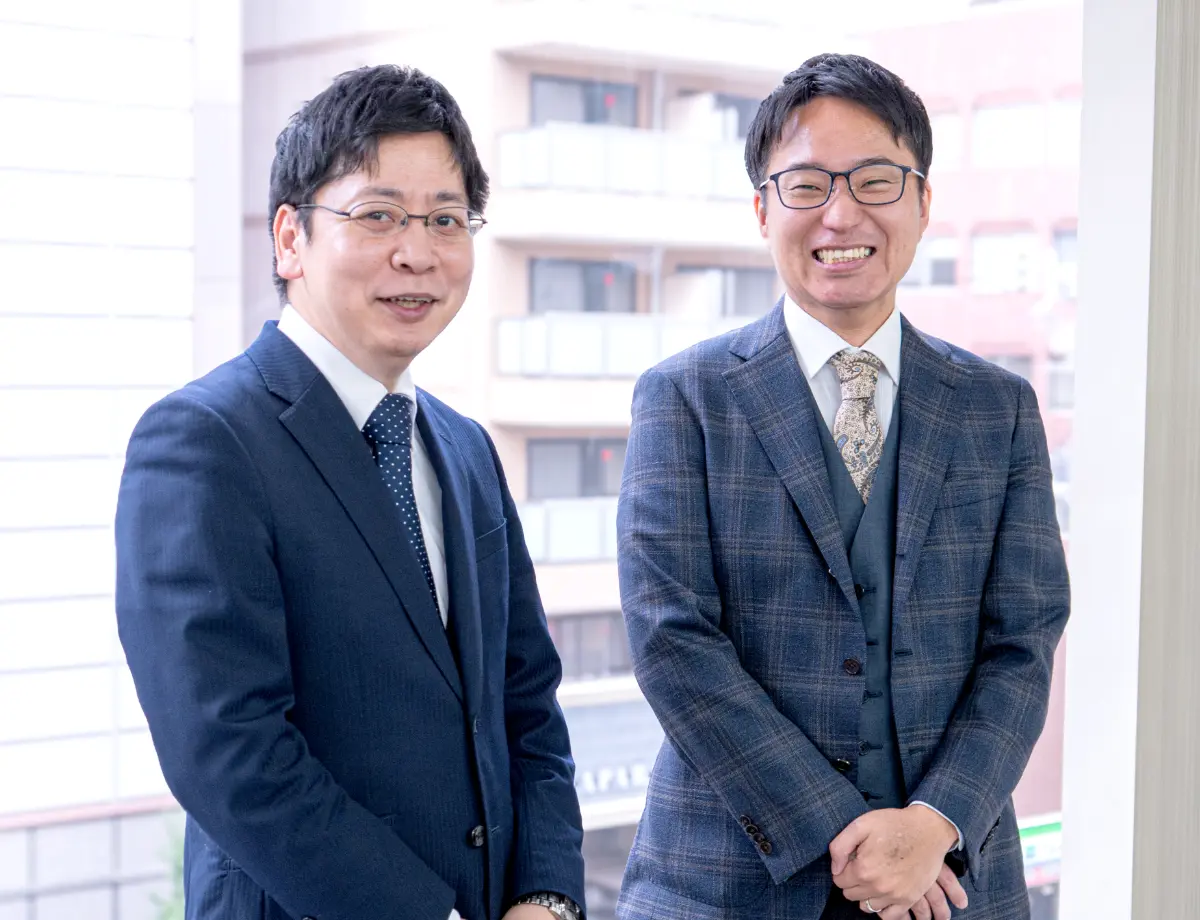 窓際で並んで笑顔を見せる、スーツ姿の二人の男性ビジネスマン