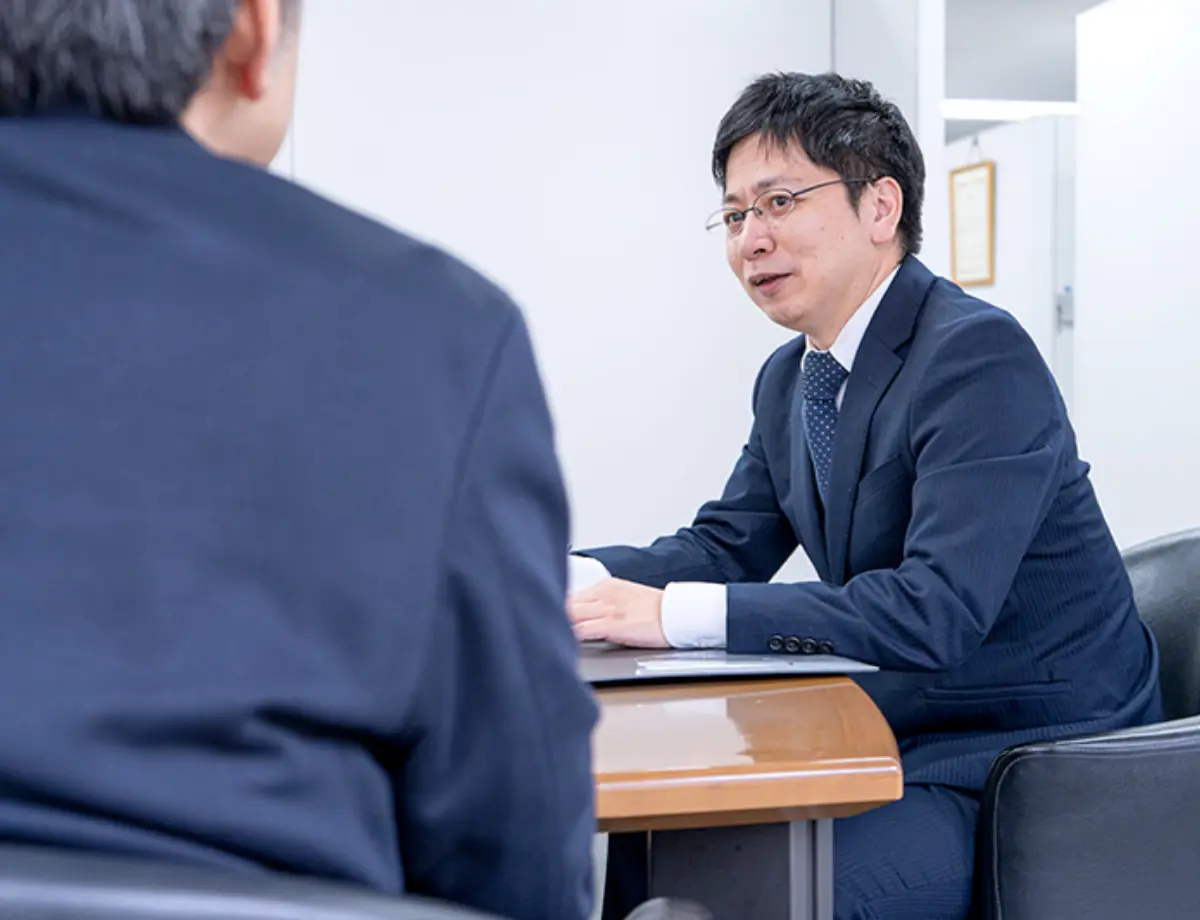 オフィス内での打ち合わせ風景。デスク越しに向かい合い、スーツ姿で話をしている様子