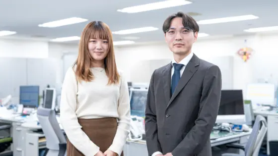 明るいオフィスを背景に並んで立つ、カジュアルな服装の女性とスーツ姿の男性スタッフ